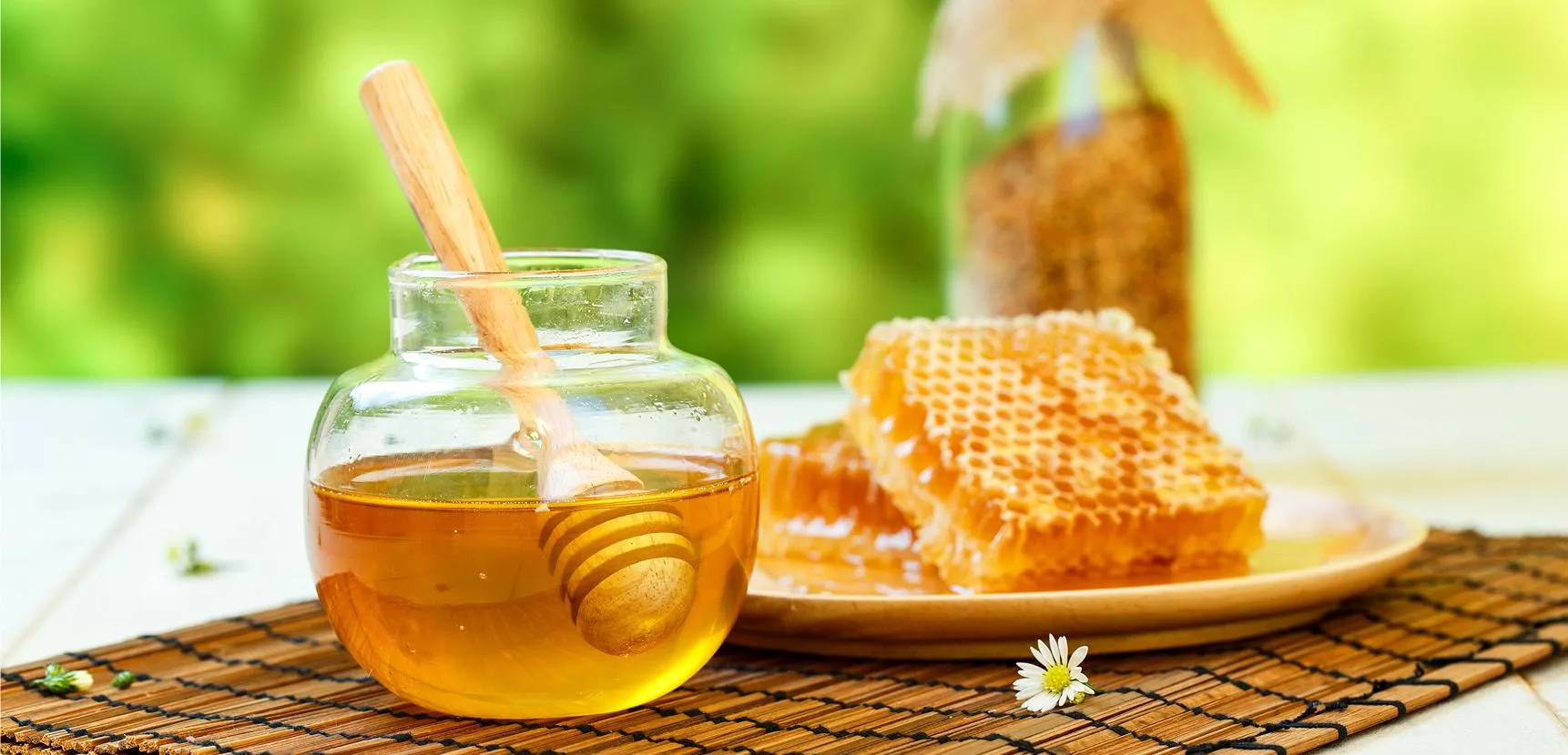 Glass jar of golden honey with dipper and stacked honeycomb on a woven mat outdoors